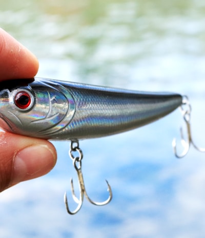 A hand holding a shiny fishing lure with red eyes and two treble hooks. The background shows a blurred view of water, suggesting an outdoor setting.