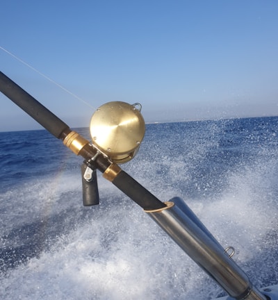 A fishing rod with a gold reel is mounted on a holder as a boat speeds over the ocean, causing splashes of water and creating a sense of motion. The sky above is clear and blue, and a rainbow is faintly visible in the spray.